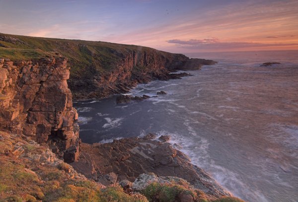 Sunlit cliffs with the sea swirling around their base. A path leads along the cliffs from a parking area near an old WWII station. Photo of Yesnaby, Orkney Islands, Scotland.