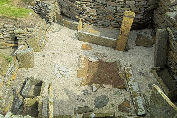 Looking down into a hut. You can clearly see the reddish hearth area, as well as slabs used to create interior partitions. Photo of Skara Brae, a prehistoric village on the Orkney Islands, Scotland.