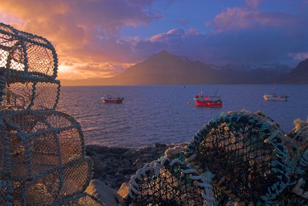 Elgol harbour at sunset. The setting sun lights up fishing boats and a stack of lobster pots. The Cuillin Hills appear in the background. Stock photo of the coast at Elgol on the Isle of Skye, Scotland. Part of the Britain Express Travel and Heritage Picture Library, Scotland collection.