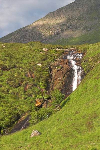 A stream tumbles down a bright green hillside, overlooked by a distant rocky hill. Stock photo of the Isle of Skye at Sconser. Part of the Britain Express Travel and Heritage Picture Library, Scotland collection.